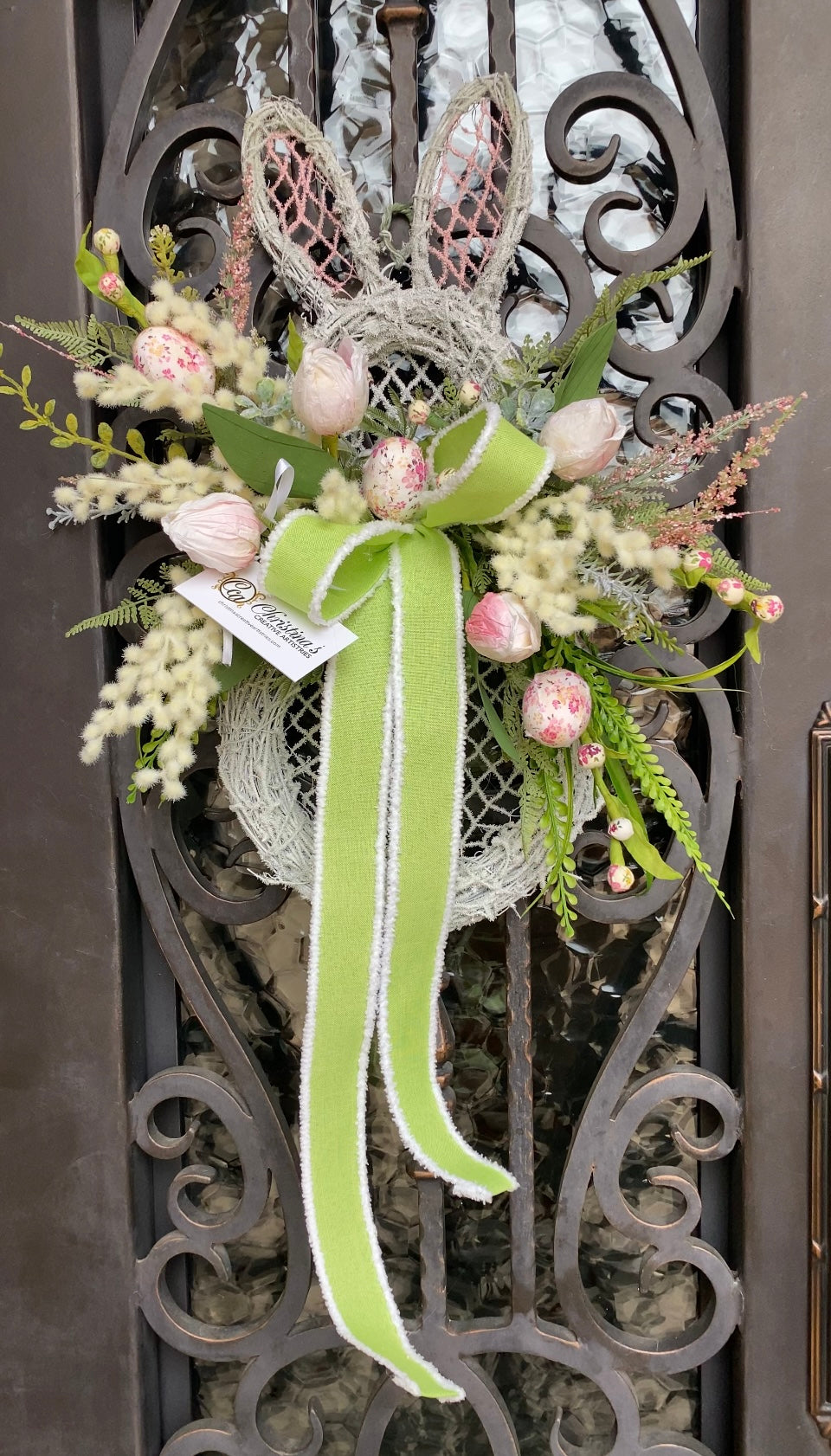 Bunny Wreath product image features an Easter wreath.   White washed bunny grapevine frame.  Bouquet with mixed greenery, fabric eggs and a beautiful , two loop bow.  Colors are white, light pink and lime green,  Measures 32'H X 18" W.  Hang on door under overhang.  Indoor outdoor use.  