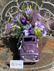 The Color Purple Truck Floral product image features the front view of the flower arrangement and truck planter. Front view gives you a closer look at the flowers, poppy pods, pampas, the hood and faux bumper lamps that are decorated with rhinestones. Indoor use or outdoors in a cover porch. Ready to ship.