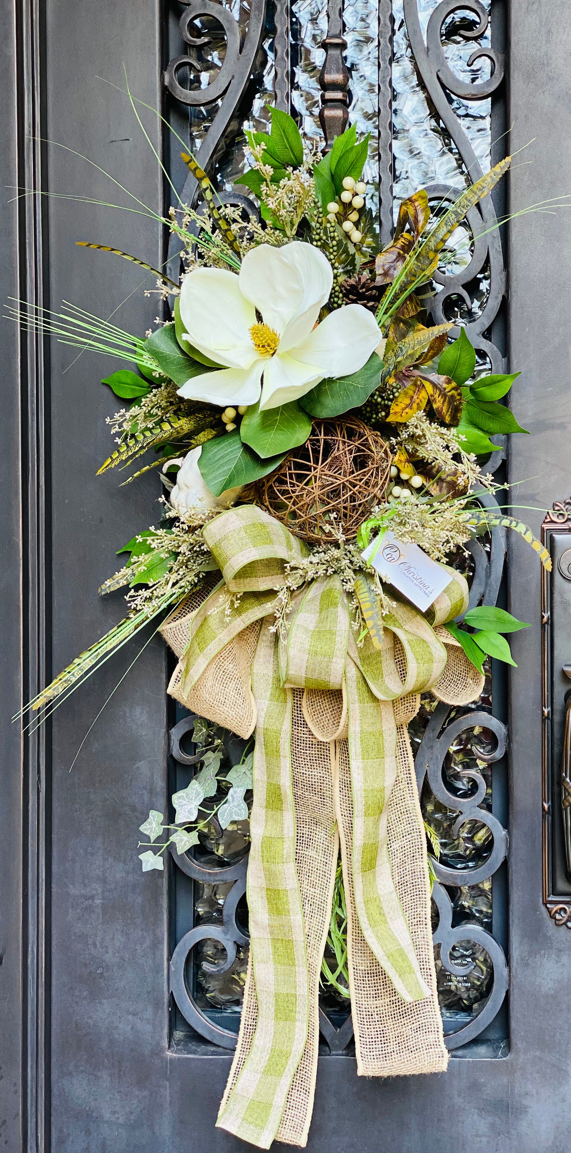 Fall Magnolia Swag product image features a beautiful fall door swag.  A large magnolia, one large, natural twig grapevine ball, one white gourd, artificial feathers, and berries with mixed greenery and a large, double bow.  Colors are cream, natural and green.  Hanging measurements are 46"L x 24"W X 12" D.  Comes ready to hang. 