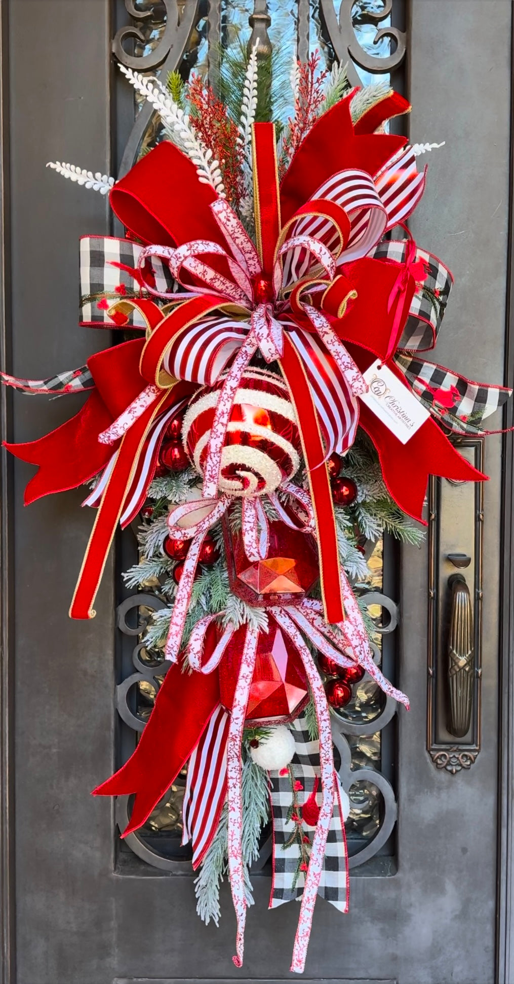 Peppermint Swirl Door Swag features a teardrop Christmas door decoration. Colors are red, white and black.  Made with bows and ornaments.  Measures 44”H X 26”W.  Indoor/ outdoor use. Handcrafted in Memphis.