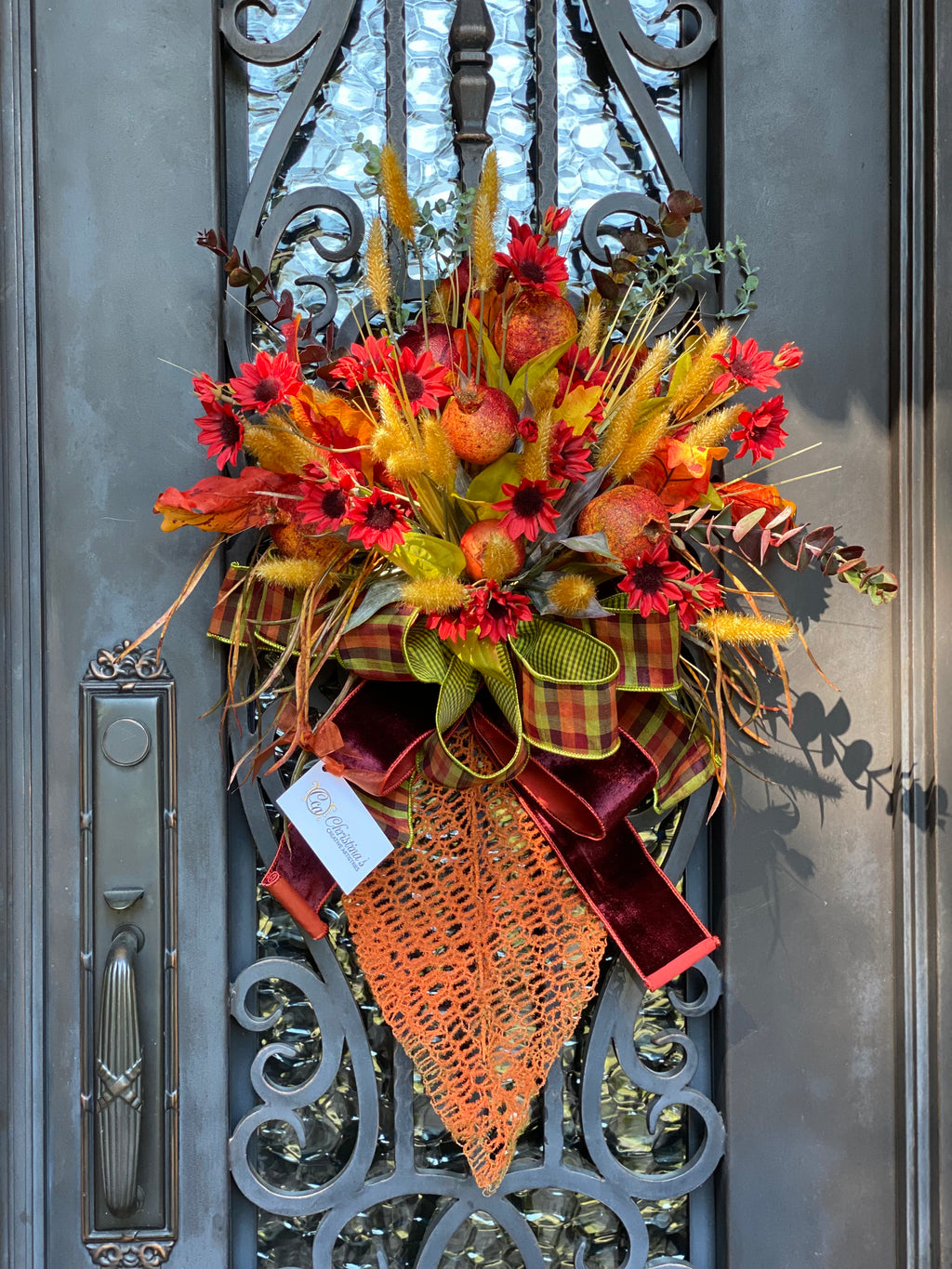 Fall Leaf Door Bouquet product image features a  fall door wreath.  Leaf wreath. Orange metal leaf, pomegranates,  gold cattails and deep orange mini sunflowers.  Large double bow with plain burnt orange and green, dark brown and burnt orange plaid ribbons.  Measures 30"H X 16"W .  Comes ready to hang.