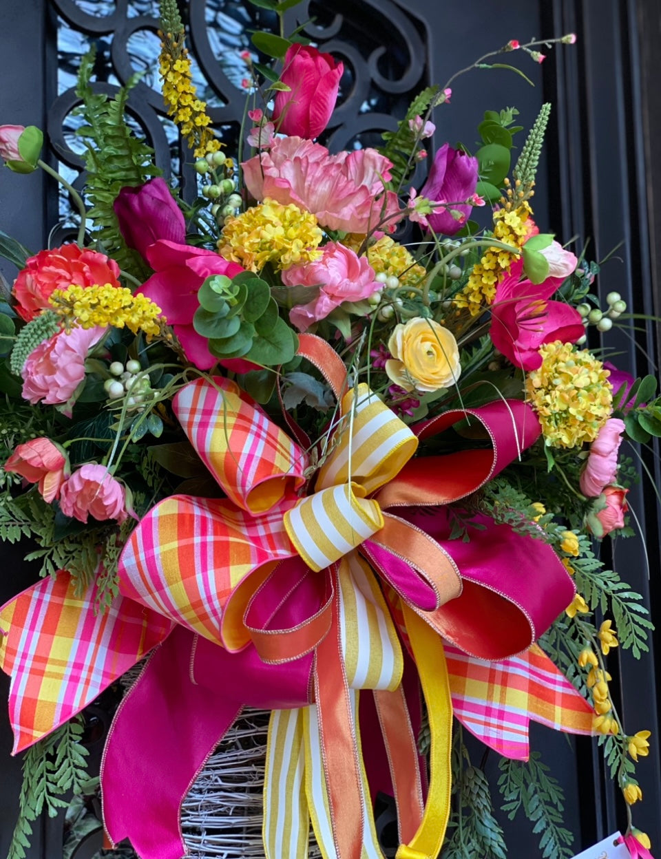 Colorful Door Basket product image features a gorgeous floral door basket.  Faux florals and greenery.  Whitewashed grapevine hanging basket.  Colors are pinks, yellow and orange in mixed greenery.  Large hand tied bow.  Measures 65"h X 28" W.  Hang on door outdoors under an overhang.