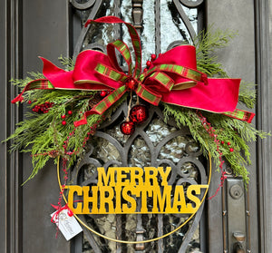 Merry Christmas Wreath product image features a Metal wreath.  Gold color.  Made with metal, greenery, fabric ribbon.  Holiday decor.  Indoor or outdoor under a covered area.  Measures 29"w X 22"H .  Comes ready to hang. 