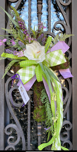 Cross Door Wreath product image features a cross wreath. Artificial. Handcrafted on a mossy wired cross frame with flowers, greenery and three ribbons. Colors are lavender, light green and white. Measures 30"H x 18"W. indoor use. Outdoor use under an overhang. Right ribbon.