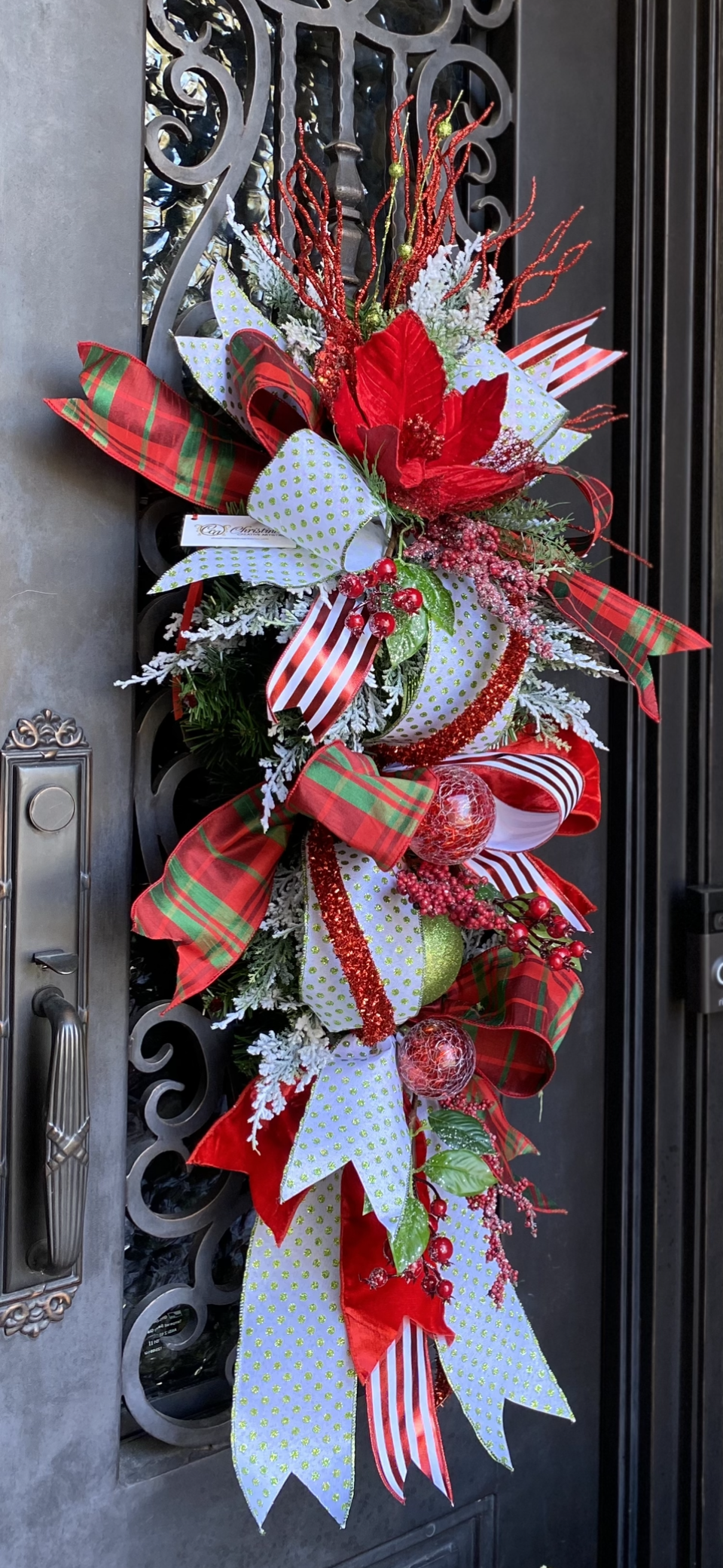 Red Green Christmas Swag product image features a Christmas door swag.  A pay on patterns.   A red and green plaid. white with green glittered polka dots and a white with red metallic striped ribbons are embellished with a red poinsettia, red, iced berries , red and green ornaments and a red glittered rope on a 30" teardrop.  Measures 50"H X 22" W X 10"L when hanging.  Hang on door with overhang. 