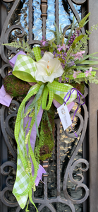 Cross Door Wreath product image features a  cross wreath.  Artificial.  Handcrafted on a mossy wired cross frame with flowers, greenery and three ribbons.  Colors are lavender, light green and white. Measures 30"H x 18"W.  indoor use.  Outdoor use under an overhang.  Left ribbon.