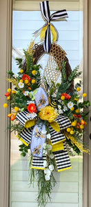 Beelicious Wreath product image features a vibrant summer wreath.  Oval grapevine frame with yellow honeycomb wire band attached.   Colors are yellow, orange, white and black and lots of greenery.   Bow is hand tied with Bee embroidered ribbon and black, white check backed with yellow