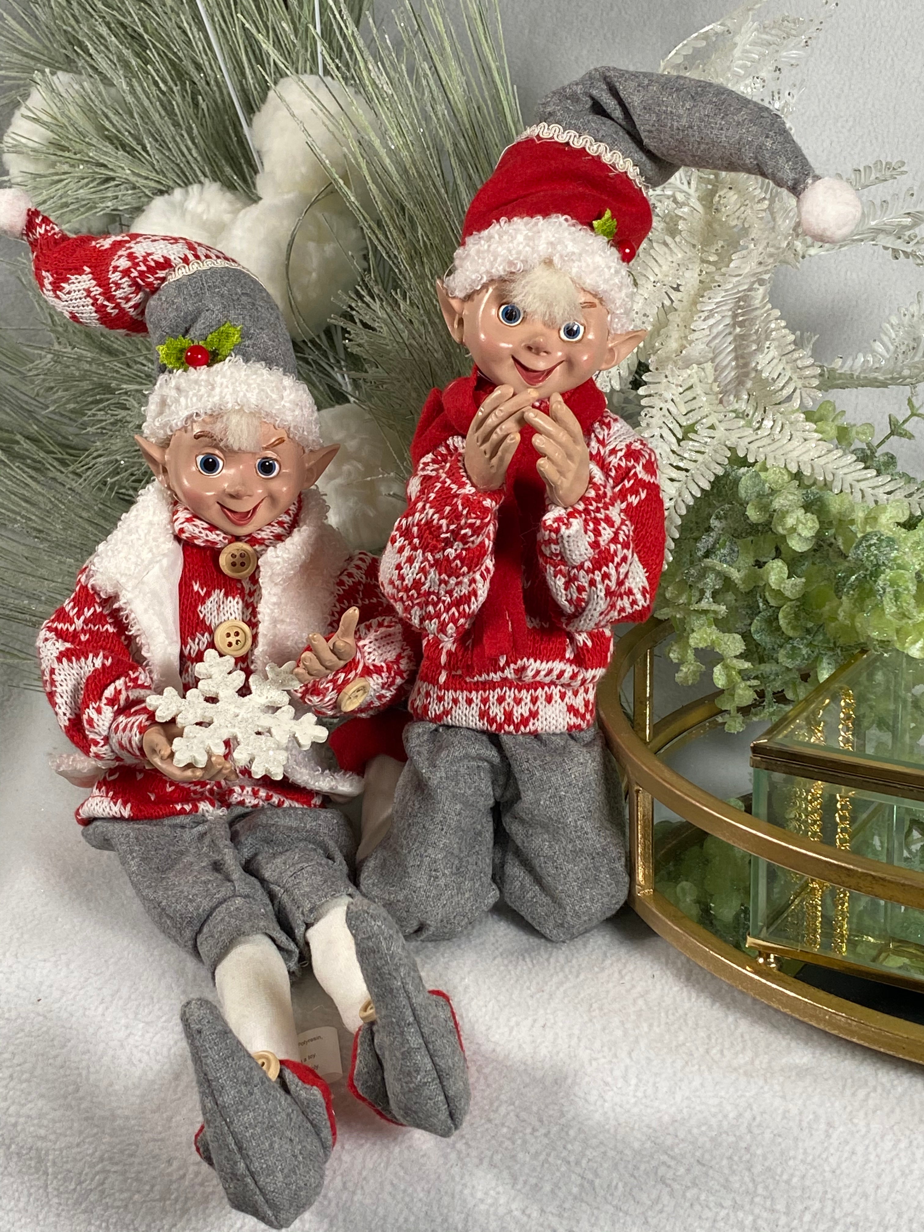 Snow Elves product image features two 16-inch, posable elves dressed warmly for play in the snow. One in a vest, the other with a scarf, both elves are dressed in warm sweaters, hats, pants and ankle high shoes. Colors are red, white and grey. Collectibles.  Posed sitting and kneeling. Decorative use only. Not intended as toys for children.