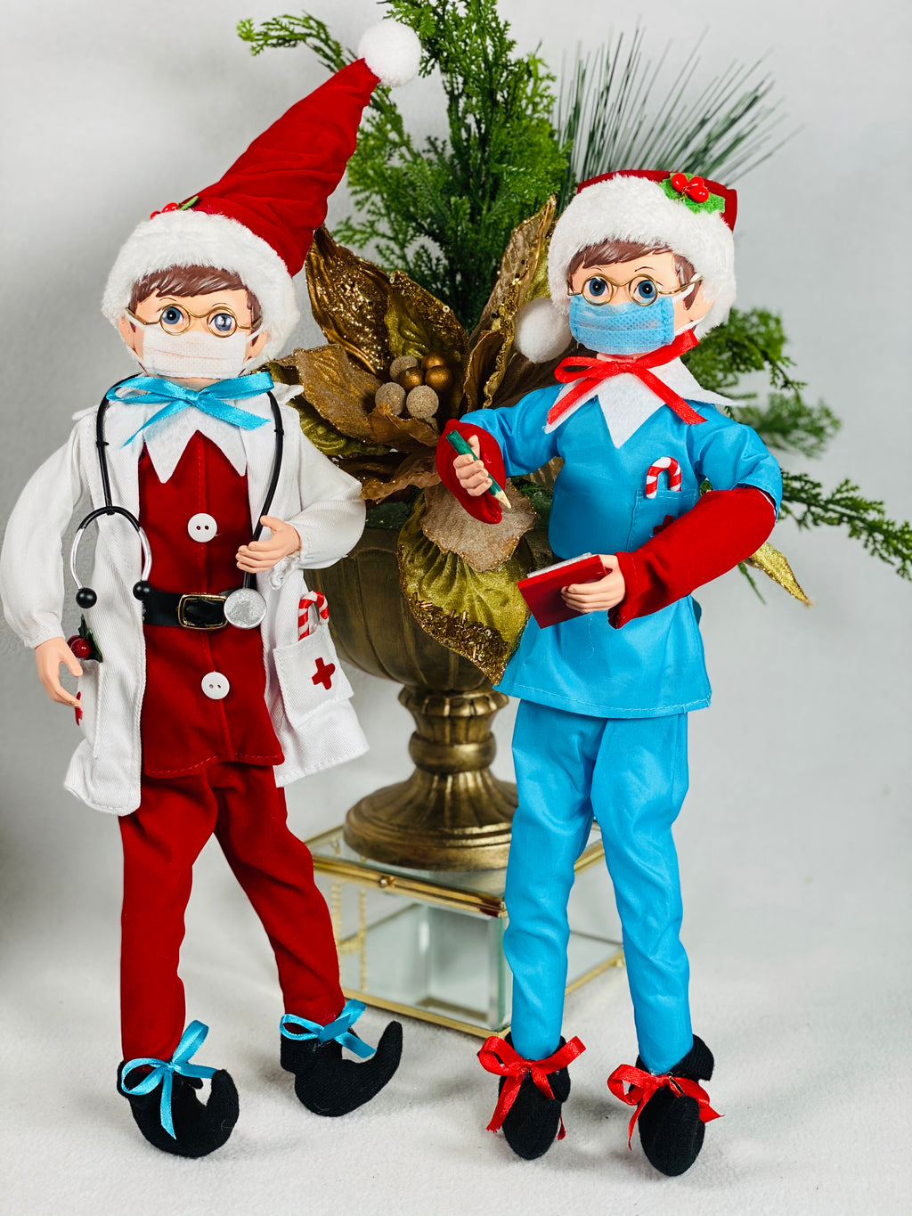 Posable Doctor And Nurse Heros Elves product image features a doctor and nurse elf.  Doctor elf has on a white mask, eye glasses, red scrubs, white coat with a stethoscope around his neck.  Nurse elf has on a blue mask, eye glasses, red shirt and blue scrubs and holding a pen and notepad.  For decorative use only.  Not intended for use as a toy for children..