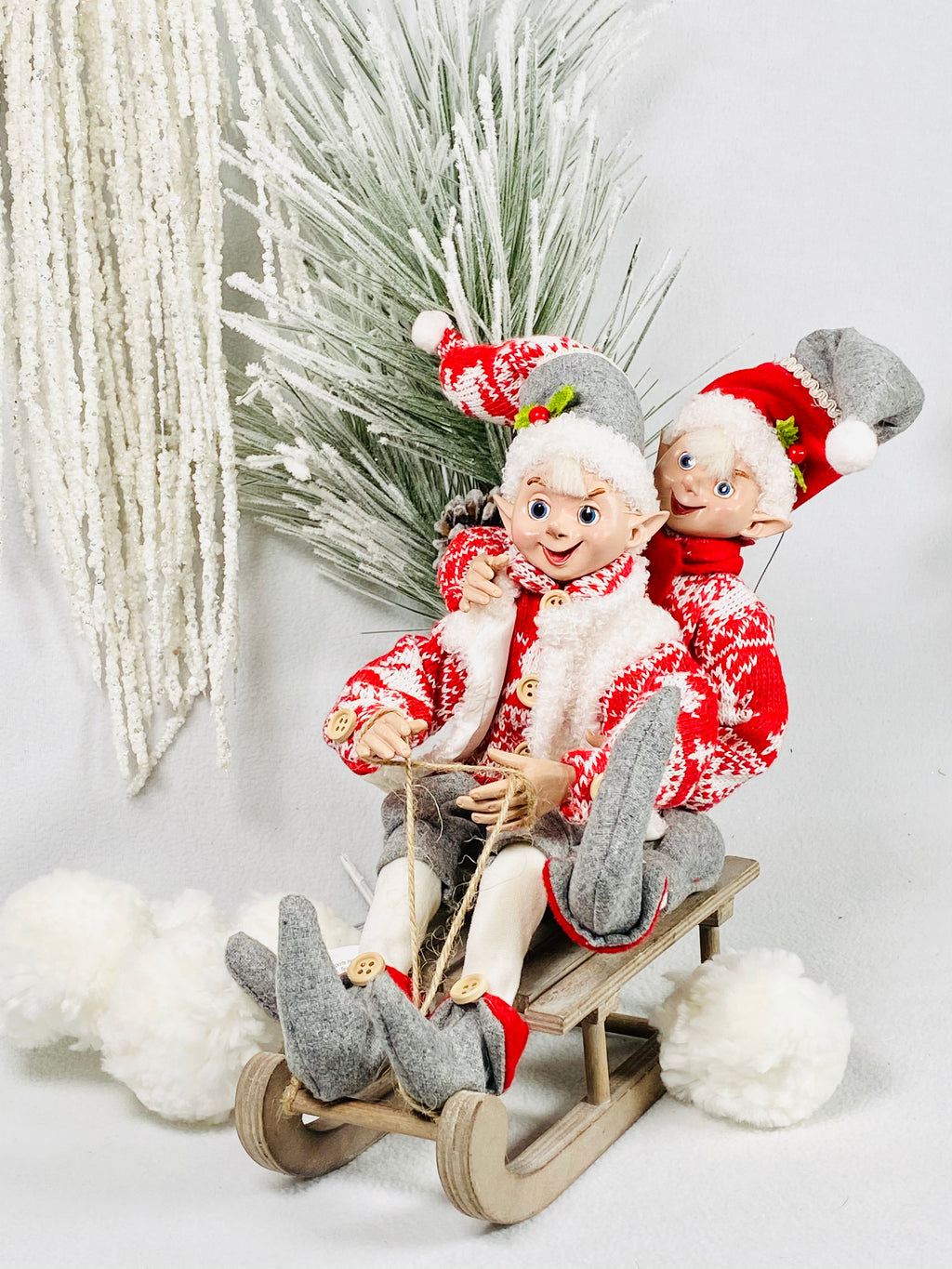 Snow Elves product image features two 16-inch,  posable elves dressed warmly for play in the snow.  One in a vest, the other with a scarf, both elves are dressed in warm sweaters, hats, pants and ankle high shoes.  Colors are red, white and grey.  Posed on a sled.  Sled not included.Collectibles.  Decorative use only. Not intended as toys for children.