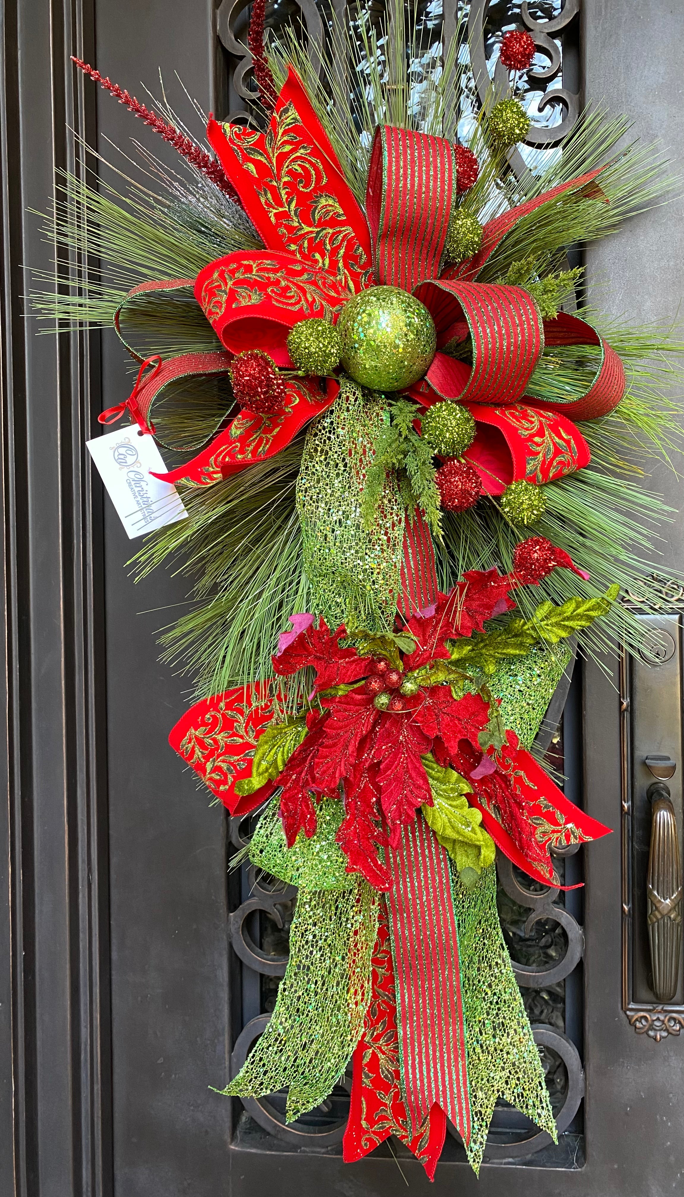 Christmas Door Swag product image features a red and green door swag. Long pine needle teardrop base. Ornaments , poinsettia and three ribbons hand tied into loops with tails. Colors are red, lime green and dark green. Measures 45"L X 24" W . Comes ready to hang on a nail or knob.