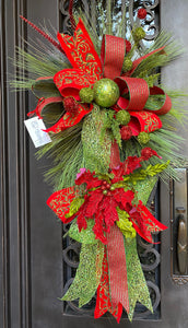 Christmas Door Swag product image features a red and green door swag. Long pine needle teardrop base. Ornaments , poinsettia and three ribbons hand tied into loops with tails. Colors are red, lime green and dark green. Measures 45"L X 24" W . Comes ready to hang on a nail or knob.