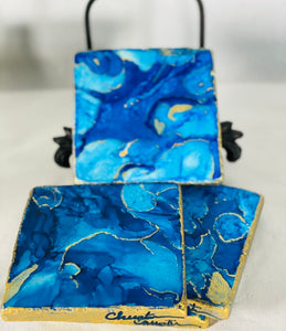 Elegant Drink Coasters product image feature a set of four stone coasters and iron stand. Hand painted on botticino marble tiles. Colors are shades of blue with gold metallic highlights. Bronze iron stand. Cork backed. signed and sealed. Measures 4" x 4". Wipe clean with a damp cloth. Ships gift wrapped for any occasion.