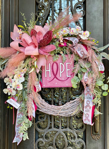 Pink Ribbon Wreath product image features a gorgeous pink wreath.   24-inch light pink grapevine frame.   Large pink velvet magnolia partially encrusted in gems, cream and light pink orchids, pink pampas and a gathering of pale green greenery.   Light pink, hot pink and white ribbon with hope, be strong and have faith inscribed.  Measures 31" H x 28" W.  Hang January till Halloween.  Comes ready to hang on a wreath hanger, nail or hook under a covered porch.
