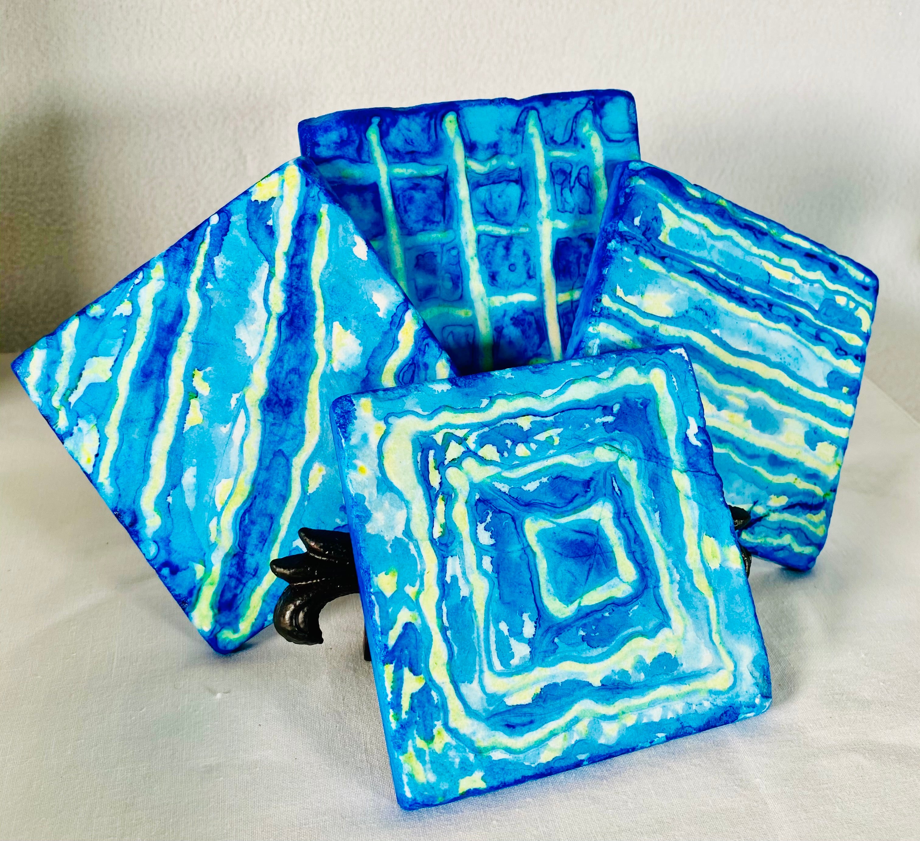 Blue Yellow Stone Coaster Set product image features a set of four stone coaters. Hand painted. Finished to last. Signed. Cork backed. Botticino marble tiles. Blue and yellow design. Wipe with damp cloth. Measures 4" X 4". Gift wrapped for Father's Day.