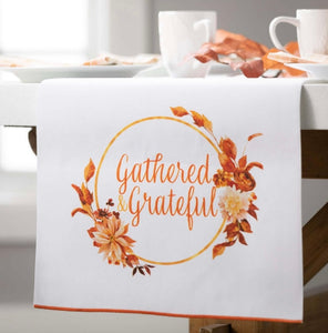 Harvest Themed Table Runner product image features The harvest -themed table runner on a table with place setting.   White cotton with Gathered Grateful script in a circle with leaves and flowers. Colors are shades of orange on white . Measures 70"L X 16.50"W.