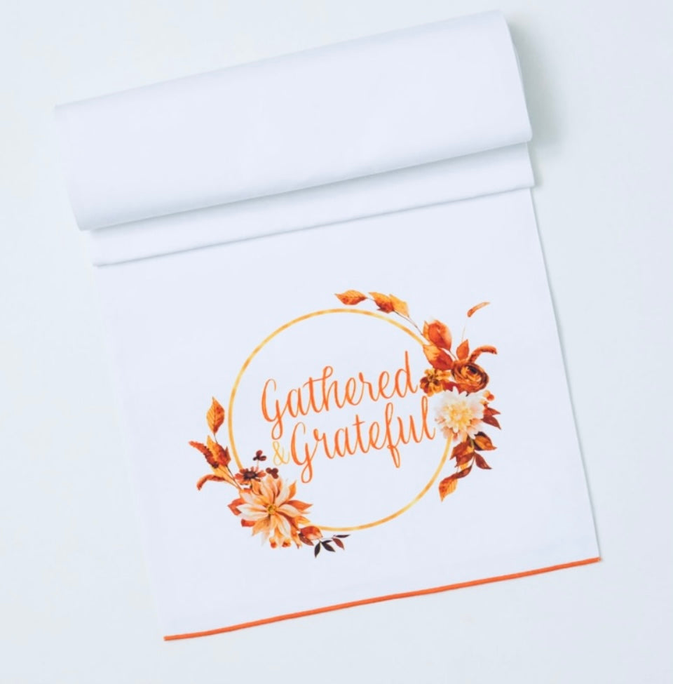 Harvest Themed Table Runner product image features  decorative textile for your table.  White cotton with  Gathered Grateful script in a circle with leaves and flowers.  Colors are shades of orange on white .  Measures 70"L X 16.50"W. 