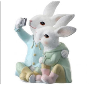 Sitting Selfie Bunnies product image features  an Easter figurine.  Two sitting bunnies taking a selfie.  White bunnies wearing  clothing in pastel hues of pink, yellow, blue and green.   Made of resin.  Measure 7" H.  Indoor use. 