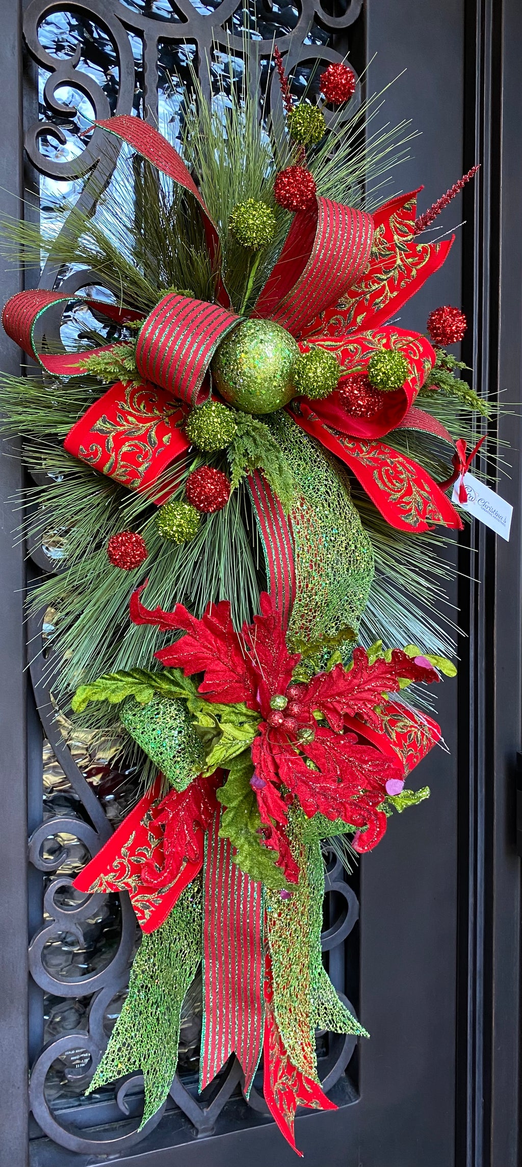 Christmas Door Swag product image features a red and green door swag.  Long pine needle teardrop base.  Ornaments , poinsettia and three ribbons hand tied into loops with tails.  Colors are red, lime green and dark green.  Measures 45"L X 24" W .  Comes ready to hang on a nail or knob. 