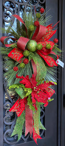 Christmas Door Swag product image features a red and green door swag.  Long pine needle teardrop base.  Ornaments , poinsettia and three ribbons hand tied into loops with tails.  Colors are red, lime green and dark green.  Measures 45"L X 24" W .  Comes ready to hang on a nail or knob. 